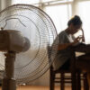 Asian unhappy woman sitting in front of working fan suffering from heat in modern house on sunny summer day in front of working fan suffering from summer heat.