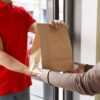 Modern delivery of any food order. Girl takes paper package from hands of courier at front door - food delivery driver