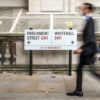 Abstract anonymous blur of a suited office worker passing a street sign indicating prominent addresses of Parliament Street and Whitehall in the UK civil service district of Westminster.
