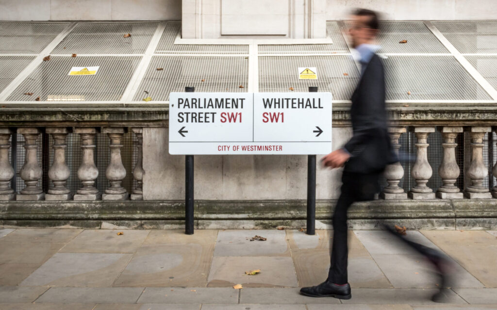 Abstract anonymous blur of a suited office worker passing a street sign indicating prominent addresses of Parliament Street and Whitehall in the UK civil service district of Westminster.
