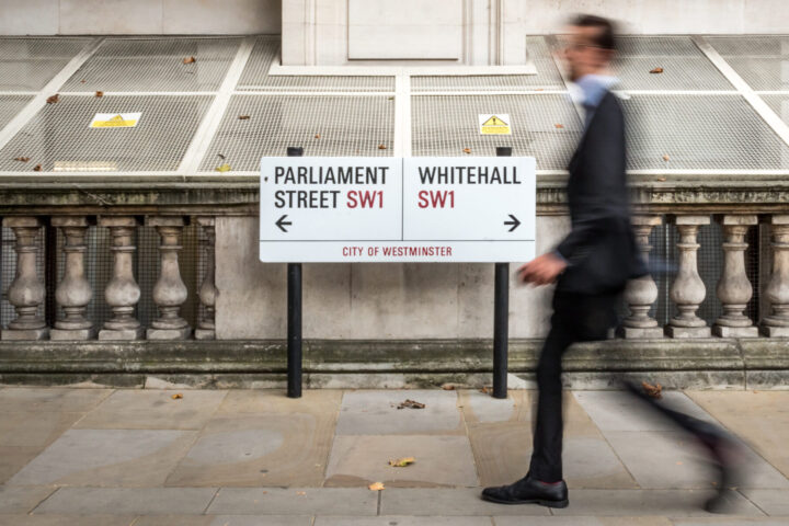 Abstract anonymous blur of a suited office worker passing a street sign indicating prominent addresses of Parliament Street and Whitehall in the UK civil service district of Westminster.