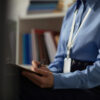 Middle aged Caucasian woman sitting and writing on clipboard, wearing identification badge, partially visible in office environment, focusing on documenting information during prisoned context