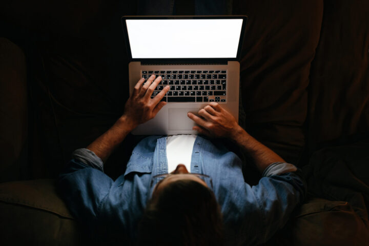 Young businessman in eyeglasses is working remotely from home late at night, looking on display, texting. Hard worker, busy man working overtime, taking work at home, has deadline. Blank screen