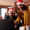 Smiling man and woman coworkers in santa hats working on laptop and celebrating xmas in office. Cheerful colleagues managing project on computer at festive decorated workplace