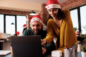 Smiling man and woman coworkers in santa hats working on laptop and celebrating xmas in office. Cheerful colleagues managing project on computer at festive decorated workplace