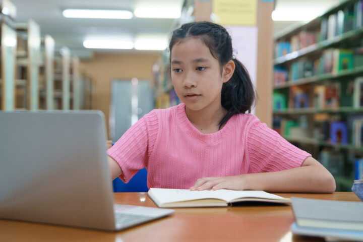 Online education, e-learning. Happy elementary school girl in stylish casual clothes, studying remotely, using a laptop, listening to online lecture, taking notes while sitting at library.